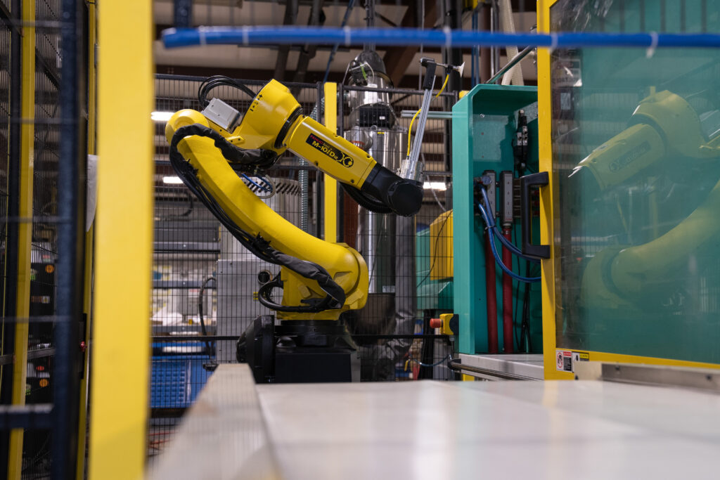 A photo of a yellow 6-axis robot at the end of a conveyor belt. It is mounted near a turquoise injection molding machine.